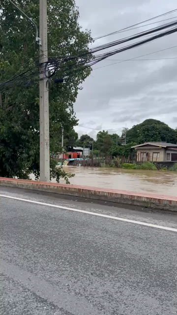 Video from the adjacent road there is a brown colored river swollen far beyond its normal level, in the distance we see pipes pumping brown water from a klong out of view, the water is almost as high as the bridge over it