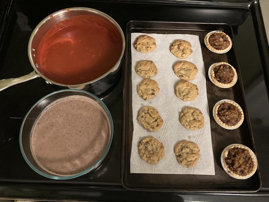 Photo taken on a black stove top with a pot of tomato soup on the top left, a glass bowl of black bean soup below it, and beside them a pan of 10 chocolate chip cookies and four green tomato mincemeat tarts.