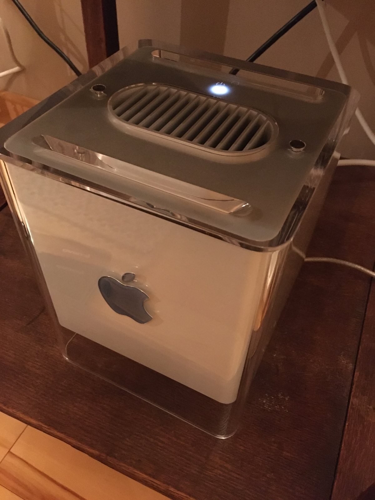 Close up of the Mac G4 cube. The top of the cube features a slot loading DVD drive, a vent for heat, and a glowing power button. the entire cube is encased in clear plastic