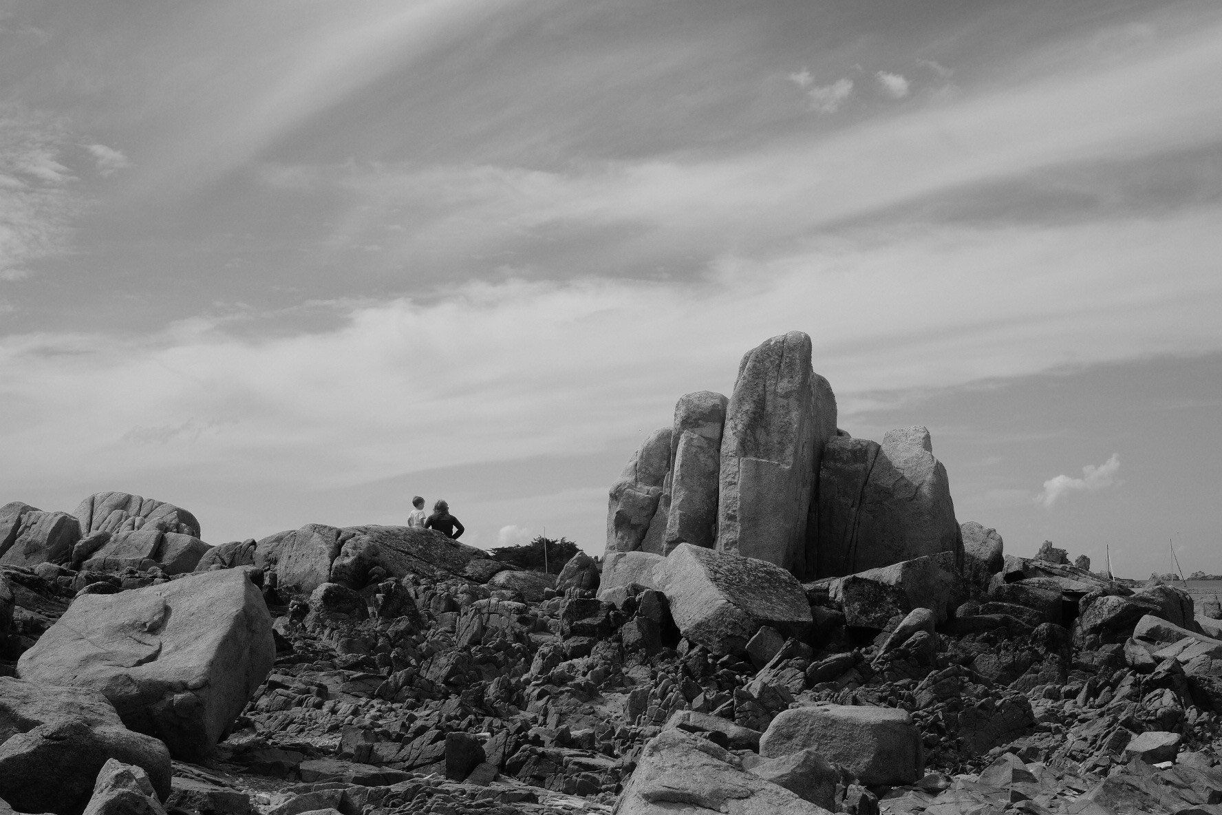 Black-and-white image. Two people sitting on a rock, on a rocky seaside, with large boulders in the background and a cloudy sky above.
