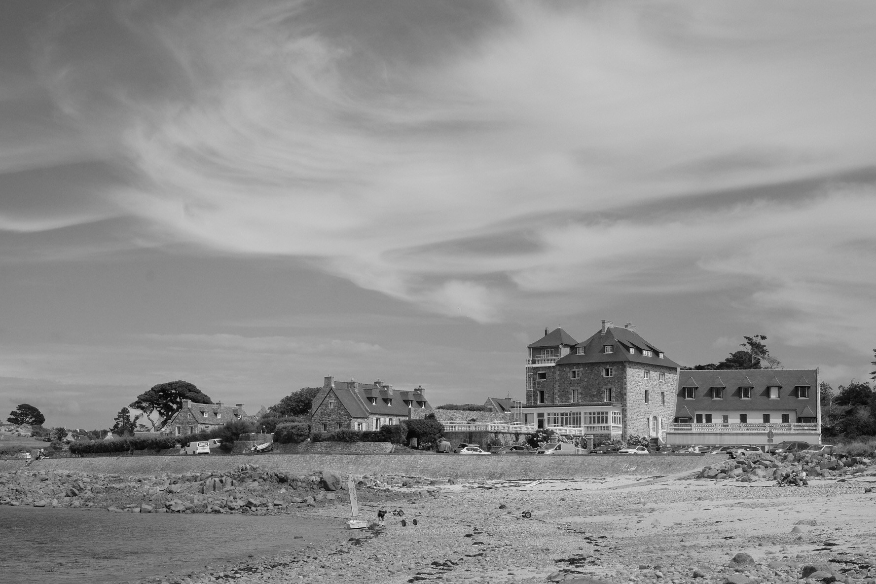 Pors Scaff, Plougrescant - French Brittany.

A coastal scene in black and white featuring a rocky shoreline and a cluster of buildings, including a prominent stone structure with a tower. Wispy clouds fill the sky, and a few people are present near the water.