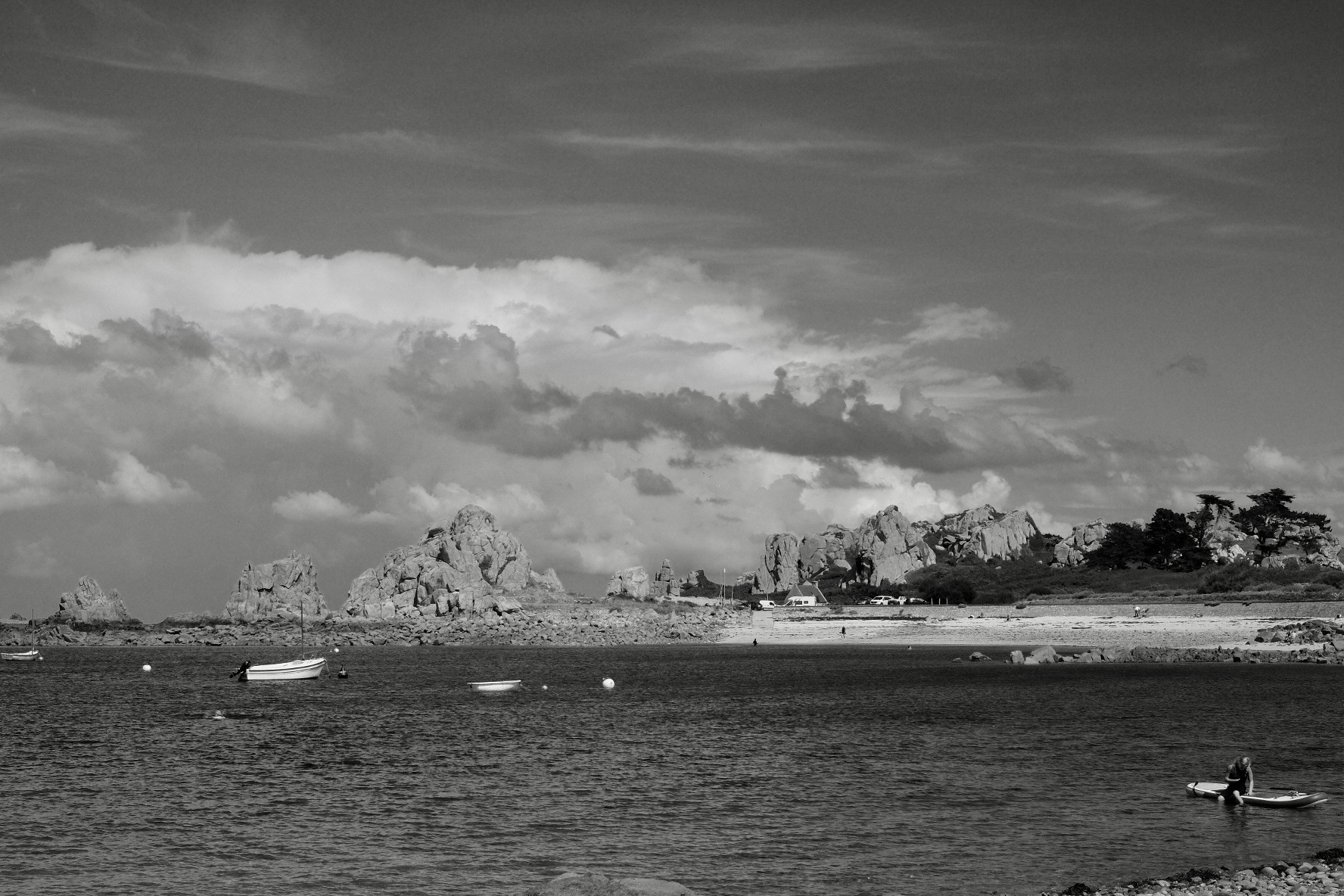 A black and white picture of seascape featuring rocky formations in the background, small boats on the water, and a person sitting on a standup paddle in the bottom right.

The rocks and partly cloudy skies are the main topics and are pretty dramatic/interesting.