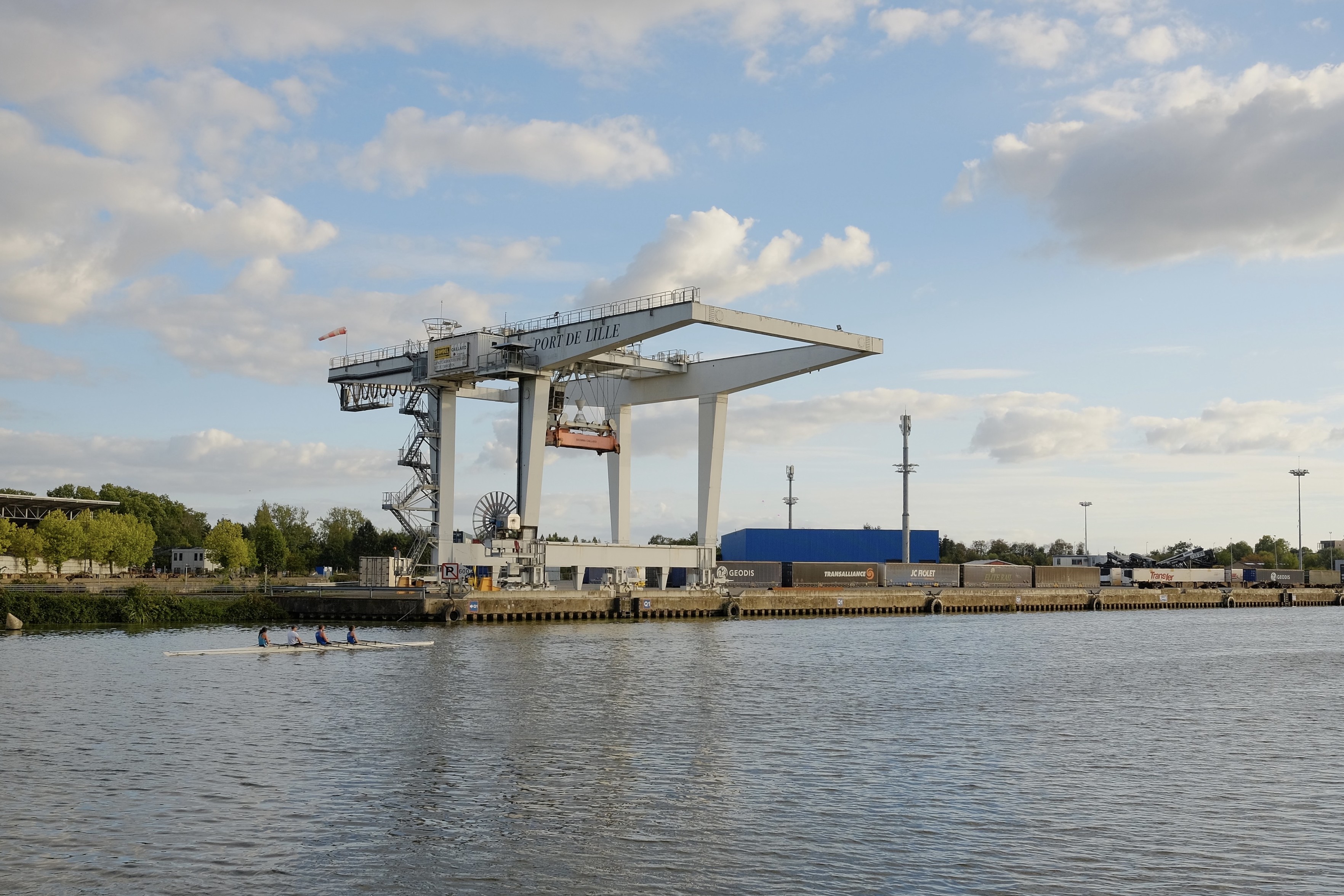 Square format, soft sunset light and colors.

A massive container crane is positioned at the Port de Lille, overlooking a calm body of water. In the foreground, a small rowing team is visible on the water, while shipping containers and structures line the waterfront behind the crane.