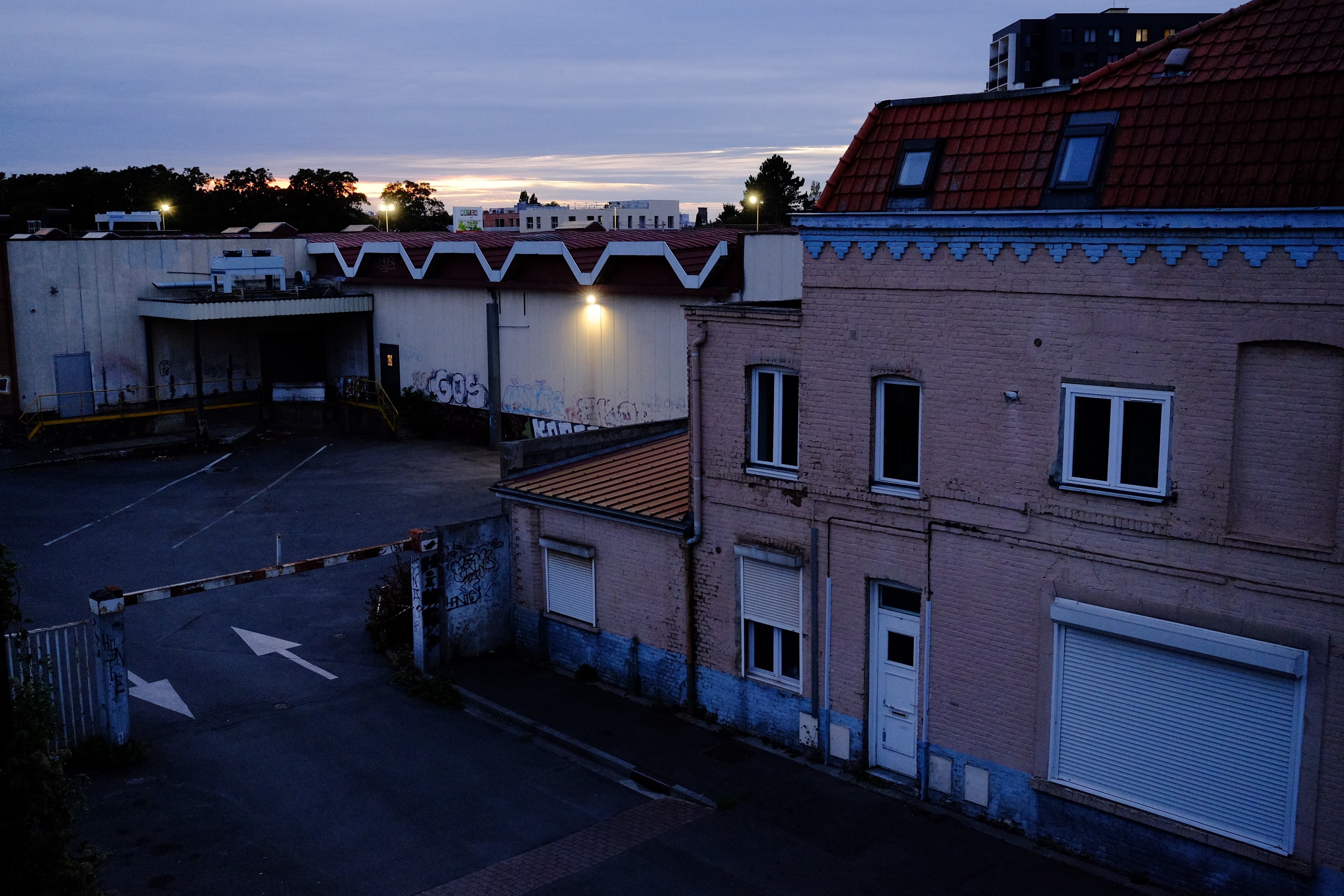 Landscape format. Urban/industrial scene.

An industrial area at dusk featuring a weathered, pink brick building with a red-tiled roof, partially closed shutters, and a parking lot. Graffiti is visible on nearby surfaces, and a loading dock with a canopy is present. 

Colors are soft and underexposed.