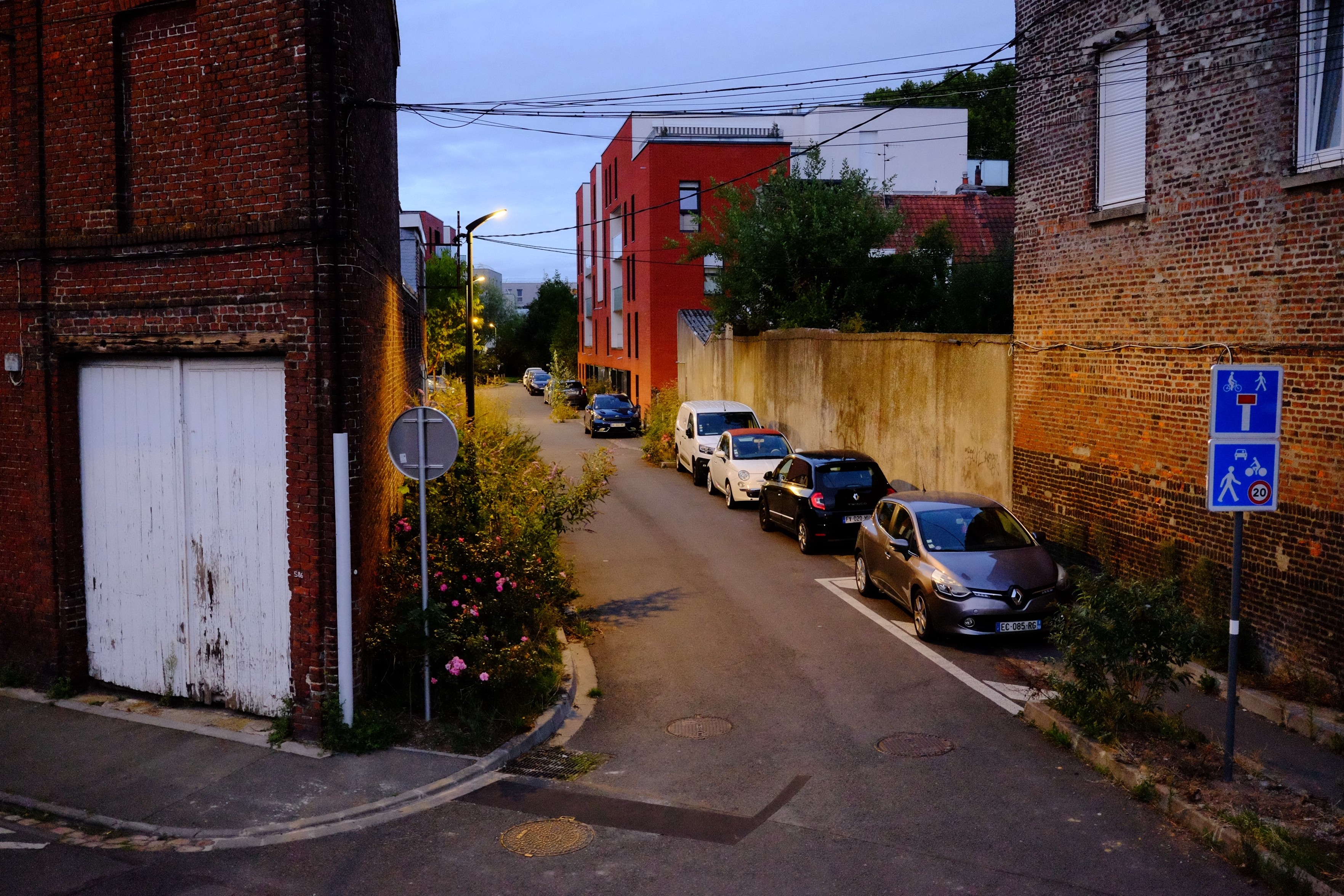 Landscape format, urban scene.

Evening street scene featuring a narrow street lined with parked cars, a brick wall, a weathered white door, and street signs indicating traffic rules. Buildings in the background include a modern red apartment structure and green foliage.

Colors are strange, dark and saturated.