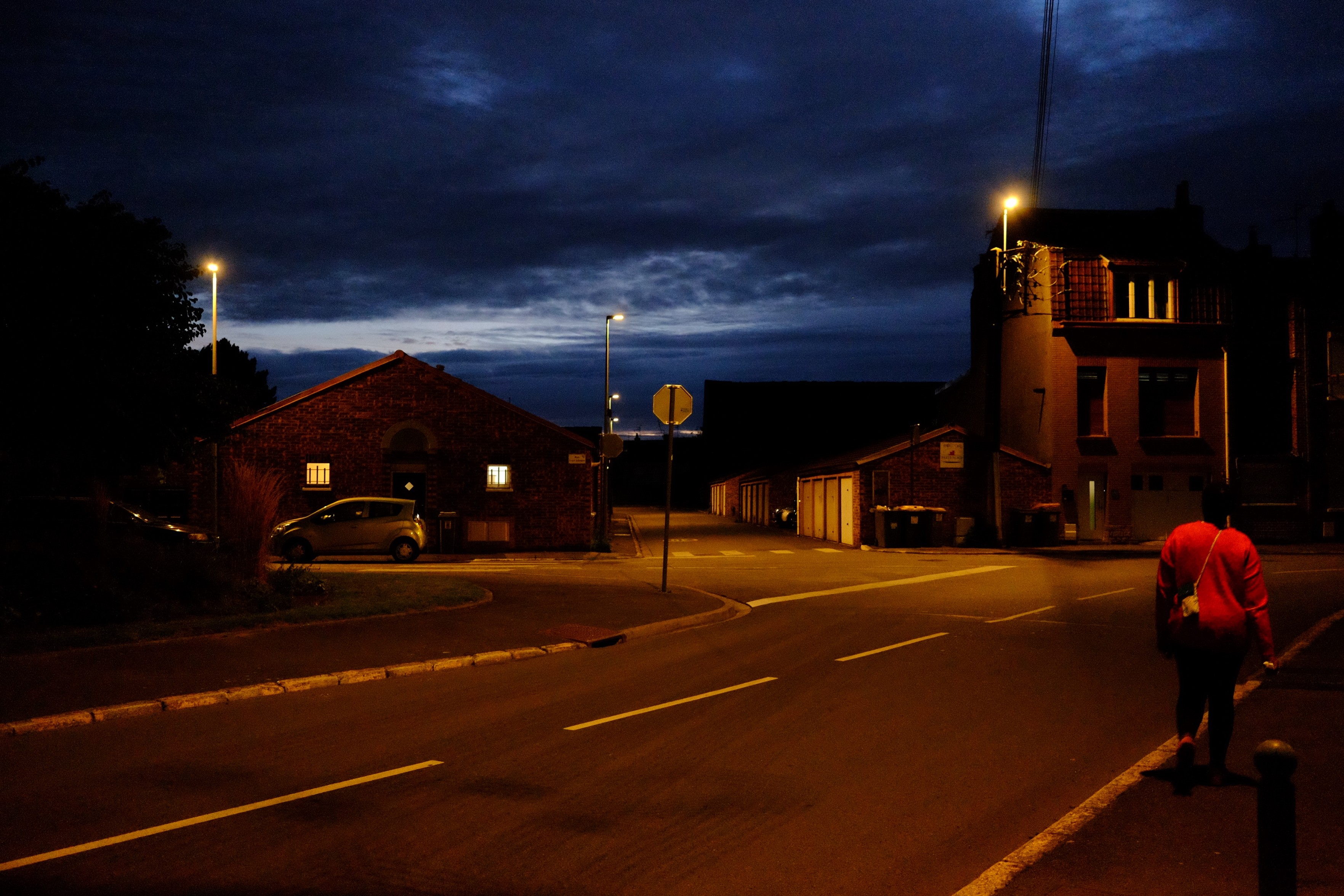 Landscape format, urban scene.

A quiet nighttime street scene with a person walking along the sidewalk. Streetlights illuminate the area, revealing a mix of buildings, including a brick structure with lit windows and garages. The sky is dark with scattered clouds.

Street has very orange/copper tones, contrasting with the dark blue skies.