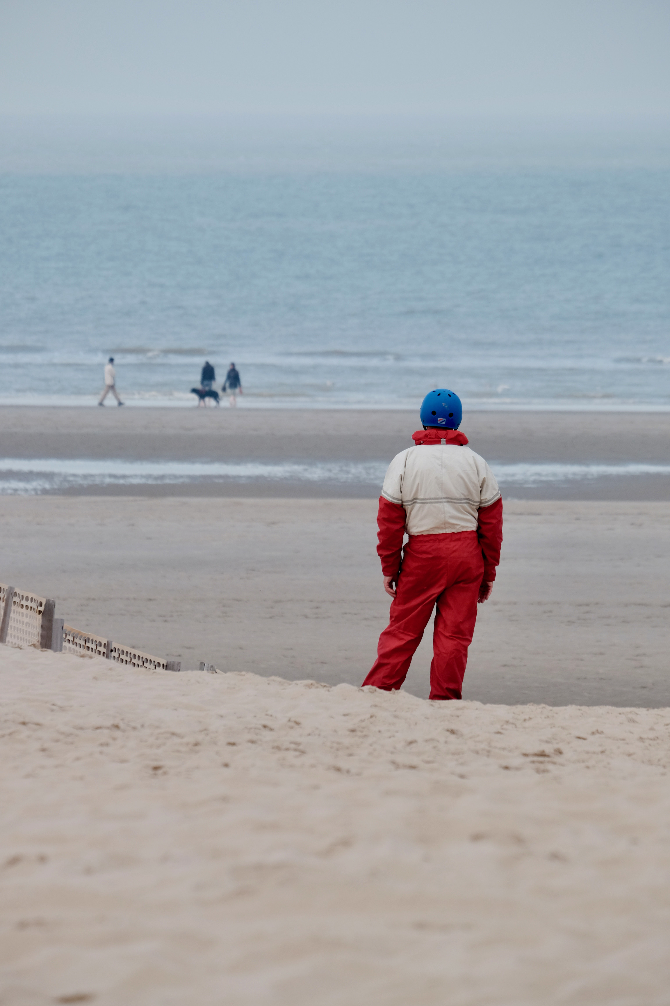 Portrait format. Beach scene.

A man in a read and beige integral suit and a contrasting blue helmet, from behind, is looking at the sea. The man's pose is a bit incongruous and seem to follow the sand beneath him.
Weather is grey/foggy and colors muted, apart the men's clothes