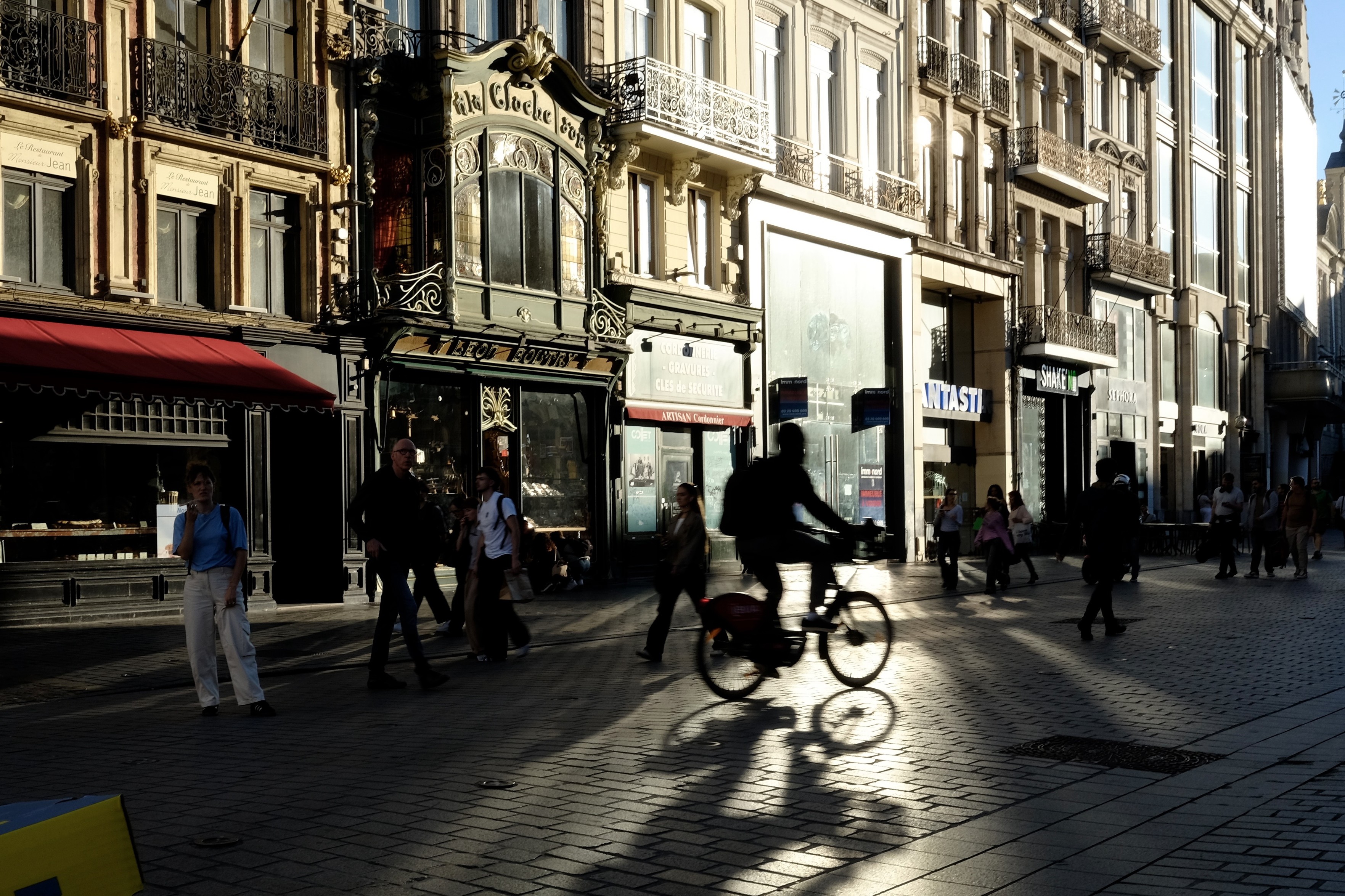 Format paysage. Scène de rue. Image sombre.
Une vitrine reflète fortement la lumière de fin d’après-midi et illumine les pavés. La silhouette sombre et floue d’un cycliste contraste avec la tache de lumière.
Façades flamandes. Centre ville.