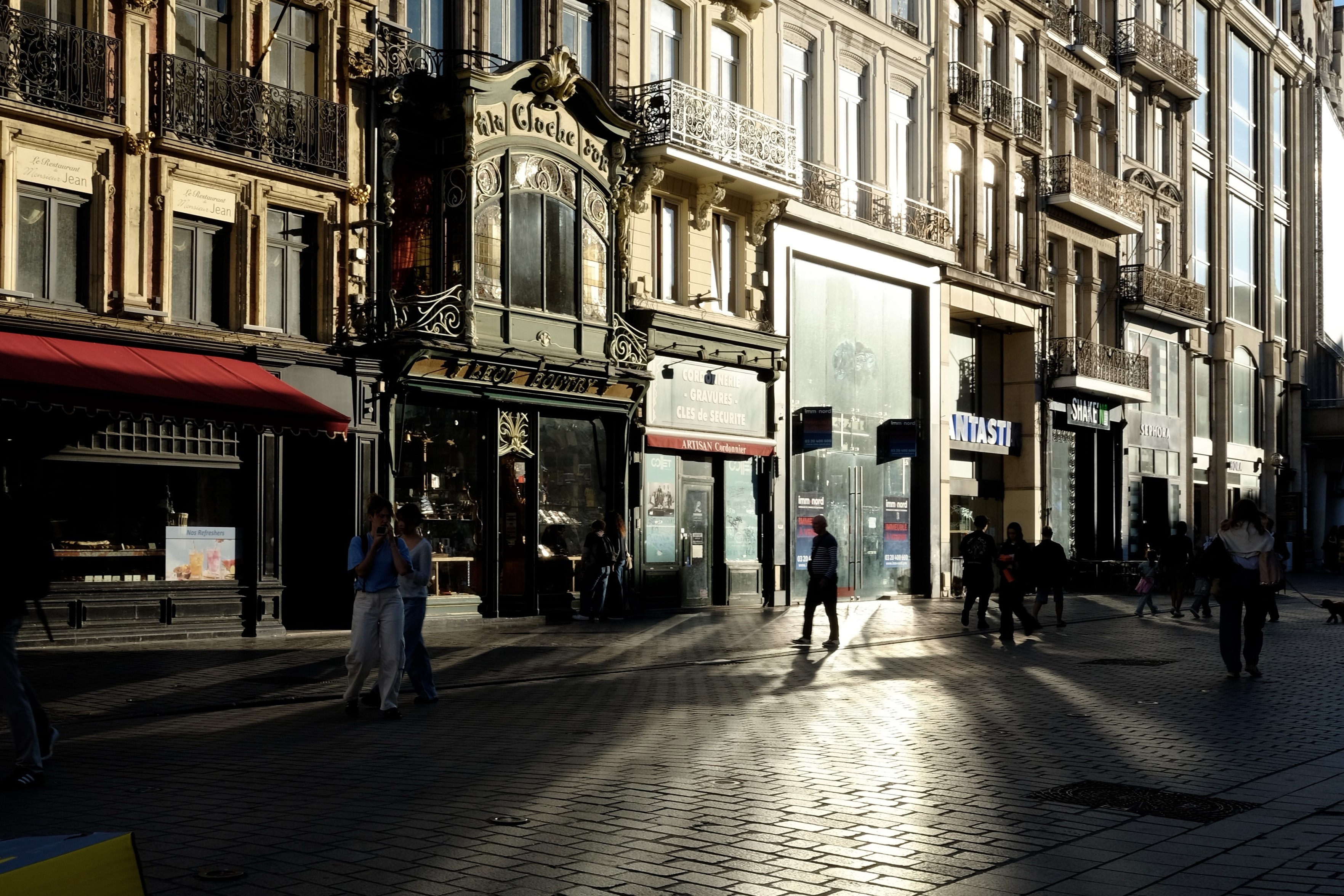 Format paysage. Scène de rue. Image sombre.
Une vitrine reflète fortement la lumière de fin d’après-midi et illumine les pavés. 
La silhouette d’un passant se découpe dans la tâche de lumière.

Façades flamandes. Centre ville.