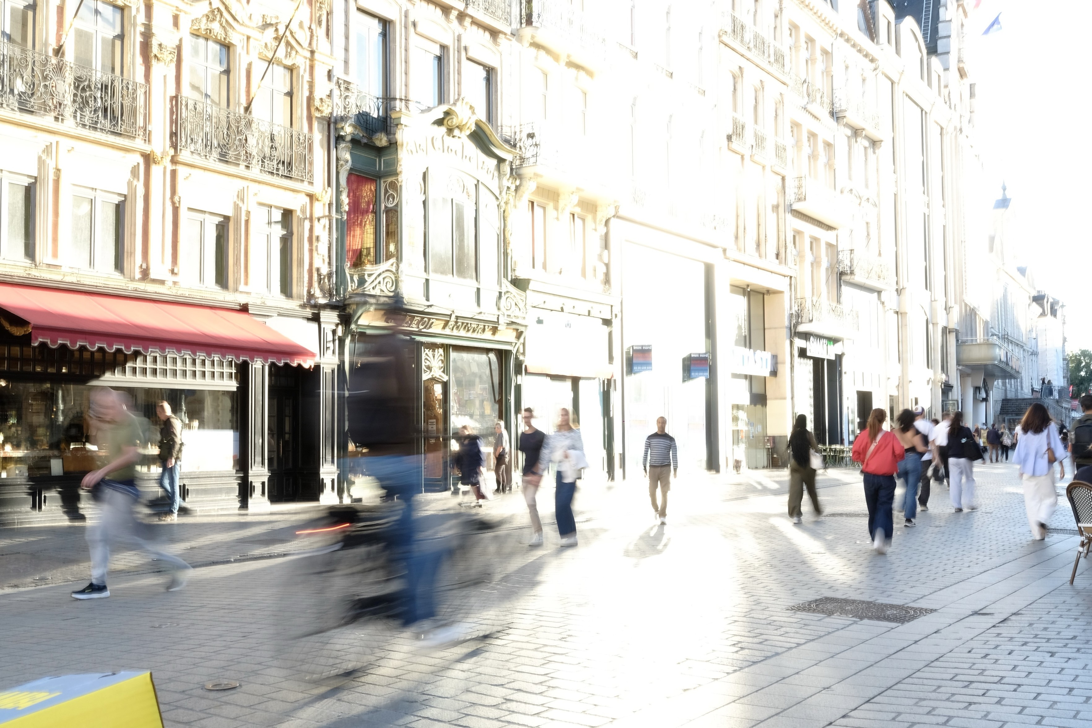 Format paysage. Scène de rue. Image claire.
Une vitrine reflète fortement la lumière de fin d’après-midi et provoque une grosse tache de lumière vers le centre de l’image. 

La silhouette d’un passant se découpe de manière frappante dans cette zone blanche.

Façades flamandes. Centre ville.