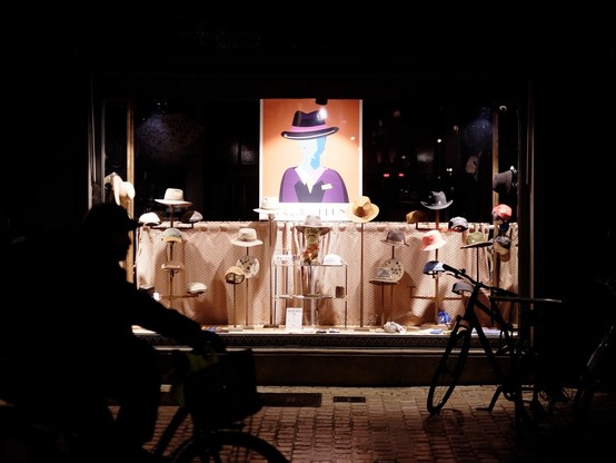 Landscape format, night scene, color.
Hat shop window displaying various hats on stands, with a colorful poster of person with a hat in the background.
A cyclist silhouette is visible in the foreground, riding past the shop from the left.
Shop window is brightly lit, the rest of scene is very dark/black.